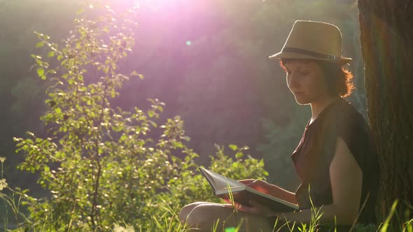 A Young Girl in a Straw Hat and Dress Sits Under a Tree, Looks Around, Then Reads a Book