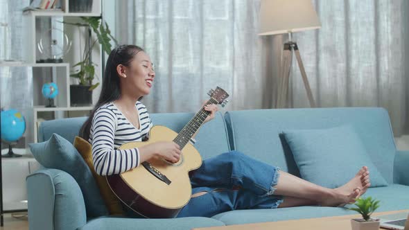 Full View Of Asian Woman Relaxing Leaning On Sofa To Sing And Play A Guitar At Home