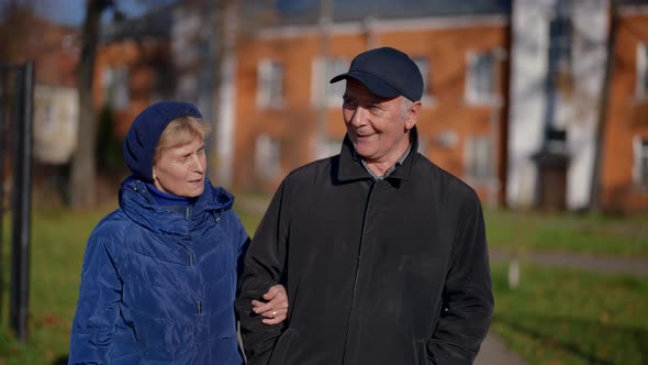 an Elderly Couple Walks Arm in Arm Against the Background of Blurred Lowrise Houses