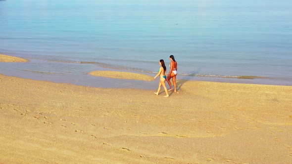 Beautiful smiling ladies on photoshoot in the sun at the beach on clean white sand and blue 4K backg