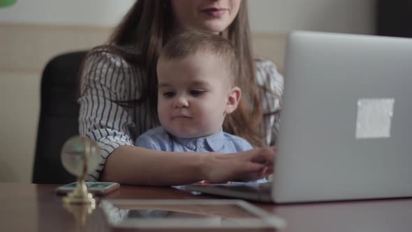 Portrait of Pretty Mother and Little Son Seated with a Laptop at the Table Indoors