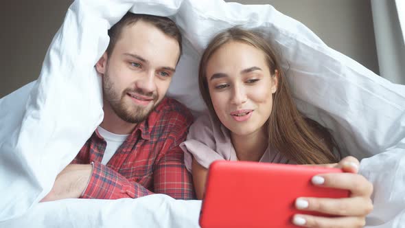 Young Couple Lying in Bed Under White Blanket Watching Video Using Cell Phone