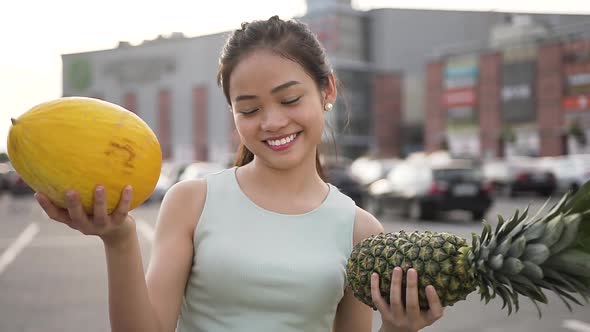Smiling Asian Girl with Melon and Peanapple in Hands Posing on Camera