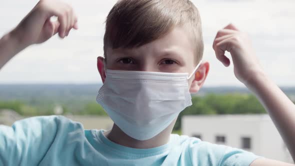 A Young Boy Puts on a Face Mask and Looks at the Camera on a Balcony in an Urban Area - Face Closeup