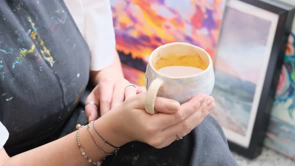 Artist Woman in Dirty Apron Resting with a Cup of Tea Sitting on the Windowsill and Looks Out the