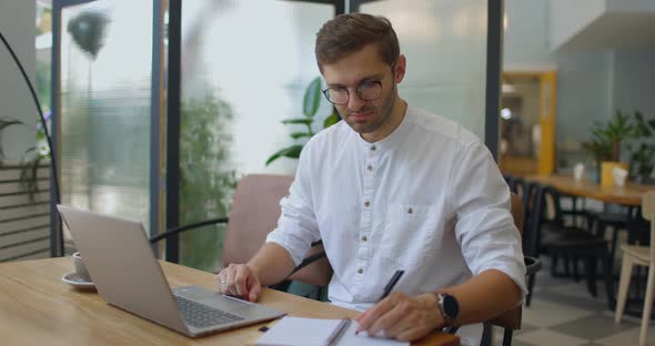 Man Having Video Call on Laptop and Making Notes in Notebook While Sitting in Small Coffee Shop