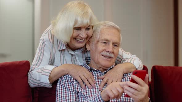 Happy Smiling Senior Couple with Mobile Phone at Home. Resting on Sofa in Cozy Living Room