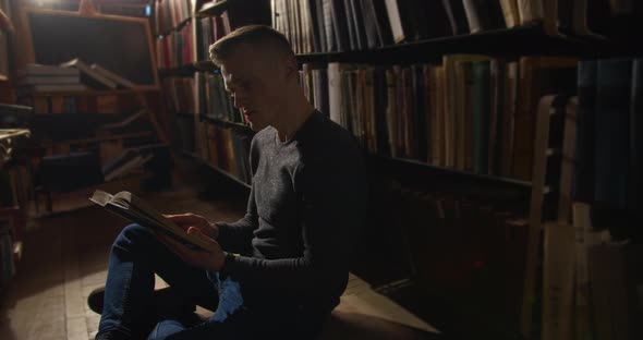 A Young Guy Is Sitting on the Floor Between the Bookshelves and Leafing Through a Book. 