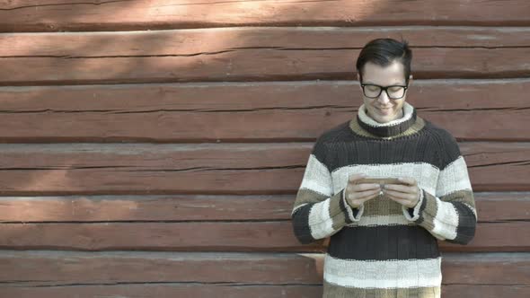 Happy Young Man with Eyeglasses Using Phone Against Wooden Wall in Autumn