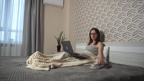 Young Girl in Glasses Lying in a Bed with a Laptop and Typing Message Than Drinks a Cup of Coffee