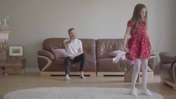 The Little Funny Girl in Red Dress Dancing in the Foreground on the Fluffy Carpet. Bearded Father