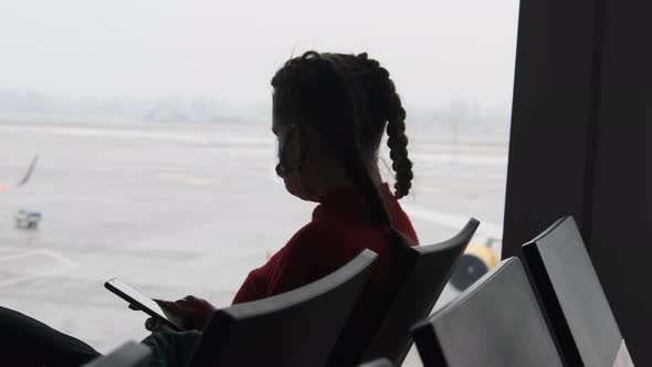 Masked Young Woman in Airport Waiting Room Sits and Using a Smartphone By Window