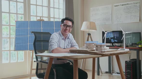 Asian Man Smile To Camera While Working With The Model Of A Small House With Solar Panel