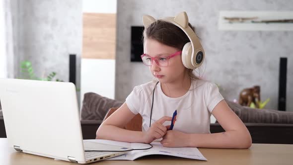 A Smiling Little Caucasian Girl with Headphones Looks at Her Laptop Sitting at the Table Listening