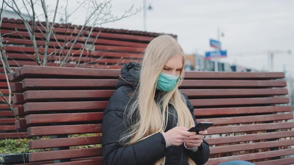 Young Female in a Medical Mask Sitting on the Bench and Using Smartphone