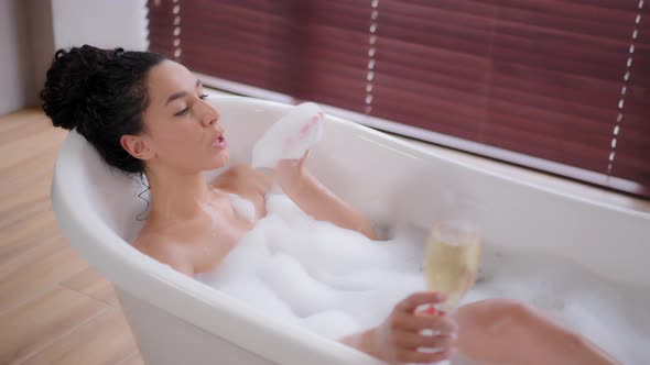 Young Playful Woman Relaxing in Hot Bath with Bubbles Holding Glass Champagne in Hand Enjoying