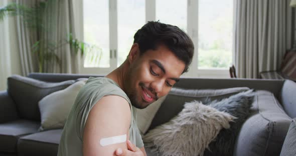 Portrait of biracial young man with bandage on arm sitting at home after immunity vaccination