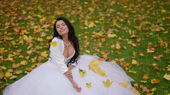 Stunning Bride is Posing on Lawn in Park in Fall Day Yellow Leaves are Falling on Her Figure
