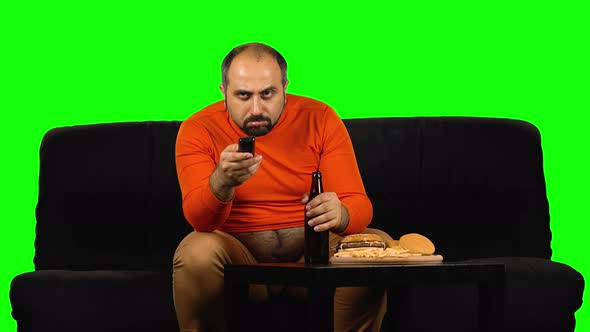 Caucasian Man with Big Belly Sits and Rejoices on Black Sofa Near Table with Fast Food, Holds Bottle