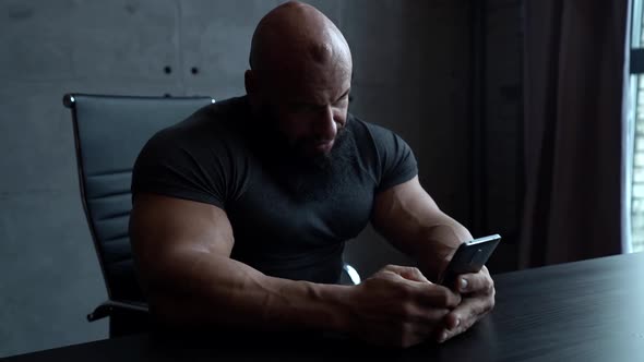 a Large Bald Man with a Beard and Big Muscles is Sitting at a Table with a Phone in Hands Against a
