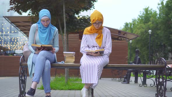 Two Muslim Students in Traditional Scarves with Books in Their Hands Are Reading Sitting on a Bench