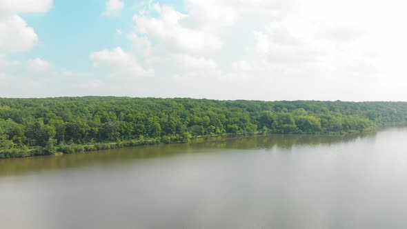 forward drone flight to a horizon and a tree line over a lake with green leaves and blue sky with wh