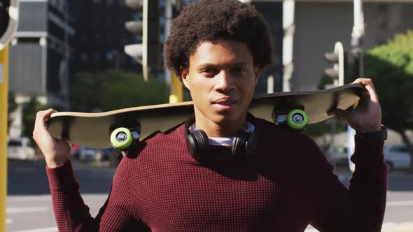 Portrait of african american man in city, standing holding skateboard on shoulders and smiling