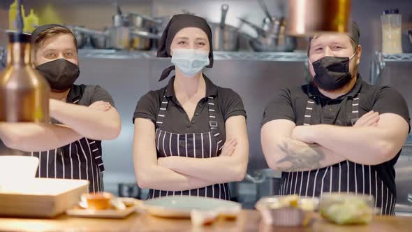 Three Confident Male and Female Cooks in Covid19 Face Masks Crossing Hands in Slow Motion Looking at