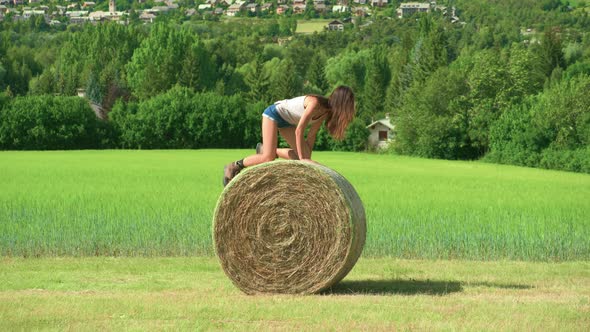 A beautiful brunette woman playing trying to roll a large bale of hay on a farm in France.