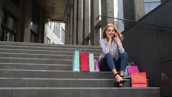 Girl Sitting on Stairs with Bags Talking on Mobile Phone About Sale in Shopping Mall in Black Friday