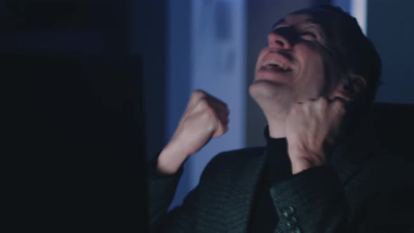 Happy Young Man Reading Great News Online Using Computer While Sitting in Dark Office Late at Night