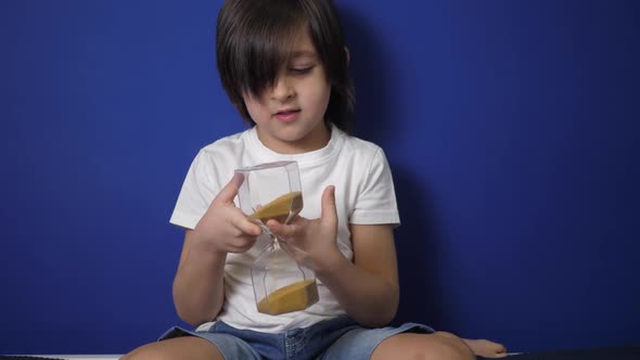 Boy Child is Sitting on a Bed Against a Blue Wall with an Hourglass