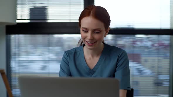 Front View of Smiling Young Female Doctor in Blue Green Medical Uniform Typing on Laptop Keyboard