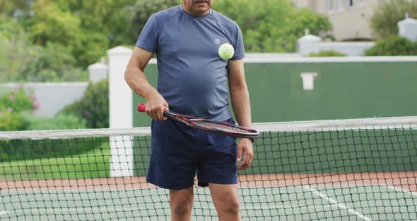 Video of happy biracial senior man holding racket and tossing ball on tennis court