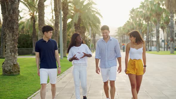 Happy Mixedrace Young Friends Communicating While Walking Together on Sunny Alley with Palm Trees