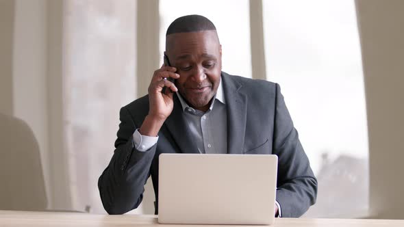 Focused African Mature Businessman Working on Laptop in Office Answering on Phone Call