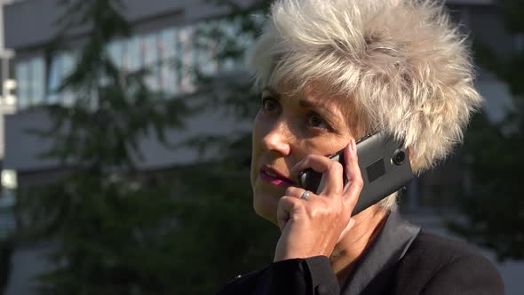 A Middle-aged Businesswoman Talks on a Smartphone in an Urban Area - Closeup - an Office Building