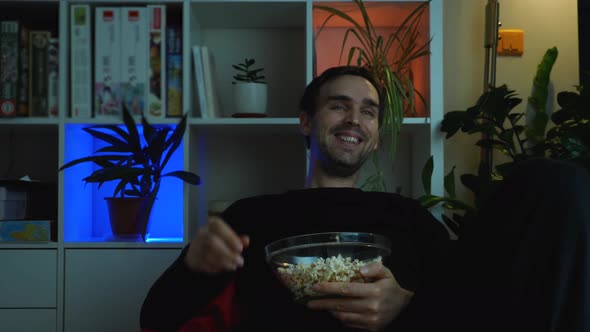 A man laughs while watching TV. A guy dressed in black sits in a sack chair with a cup of popcorn