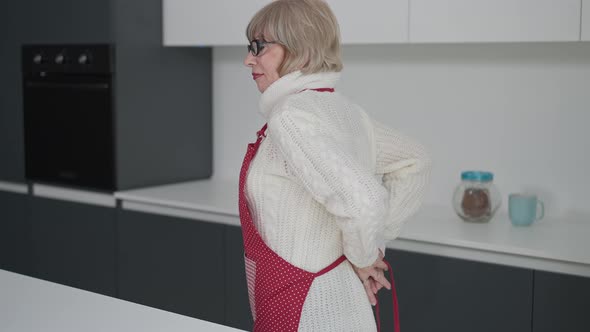 Gorgeous Senior Woman in Eyeglasses and White Sweater Tying Apron Standing in Kitchen at Home