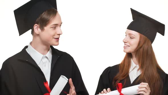 Two Caucasian Young Graduate Boy and Girl in Black Robes and Square Academical Caps Holding Scrolls