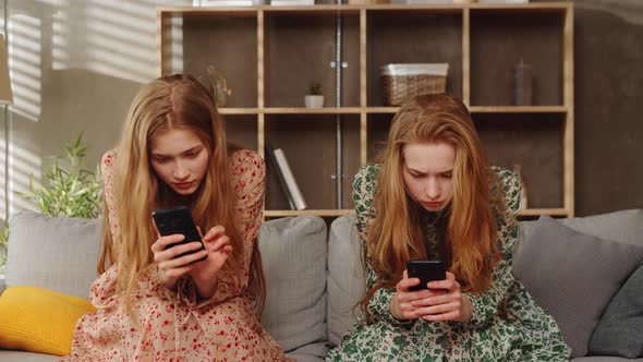 Two Young Twins Sisters Use Smartphones on Sofa in the Living Room