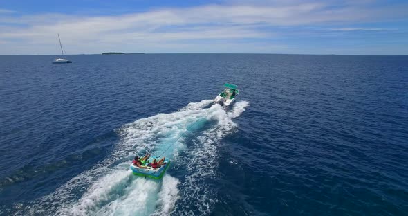 Aerial drone view of man and woman on an inflatable tube towing behind a boat to a tropical island