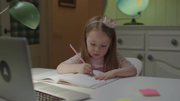 Close Up of Cute Preschool Girl Writing Down in Notebook with Pencil Looking at Laptop
