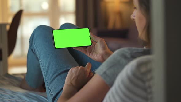 Woman at Home Lying on a Sofa and Using Smartphone with Green Mock-up Screen in Horizontal Mode