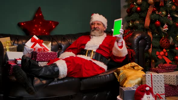 Santa Claus Lies on the Sofa Near Decorated Christmas Tree and Gift Boxes Shows Tablet with a Green