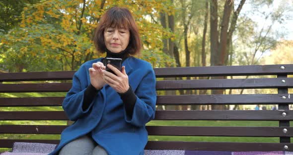 Senior Woman Is Chatting with Friends in a Smartphone While Sitting in a Cozy Park. Happy Old Woman