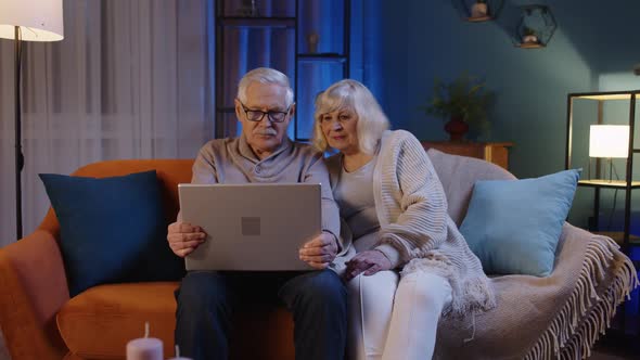 Happy Senior Elderly Couple Using Laptop Pc Talking Together Doing Online Shopping at Home Sofa