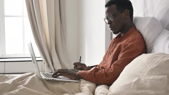 AfroAmerican Man with Glasses Makes Notes Working on Laptop