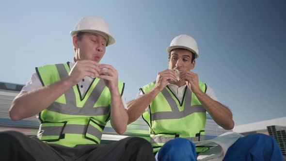 Front View of Two Hungry Warehouse Workers Enjoying Tasty Sandwiches Sitting Outdoors in Sunshine