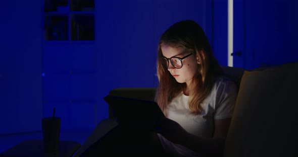Young Girl Sitting on a Sofa Using a Tablet at Night Browsing the Internet Before Pausing To Check